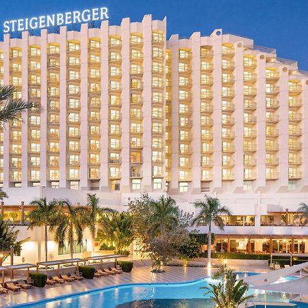 Steigenberger Hotel & Nelson Village, Taba Exterior foto The image shows a large hotel building with multiple floors, featuring a modern design. The hotel is illuminated in the evening, giving it a warm glow. In front, there is a swimming pool surrounded by palm trees and lounge chairs. The name "Steigenbe