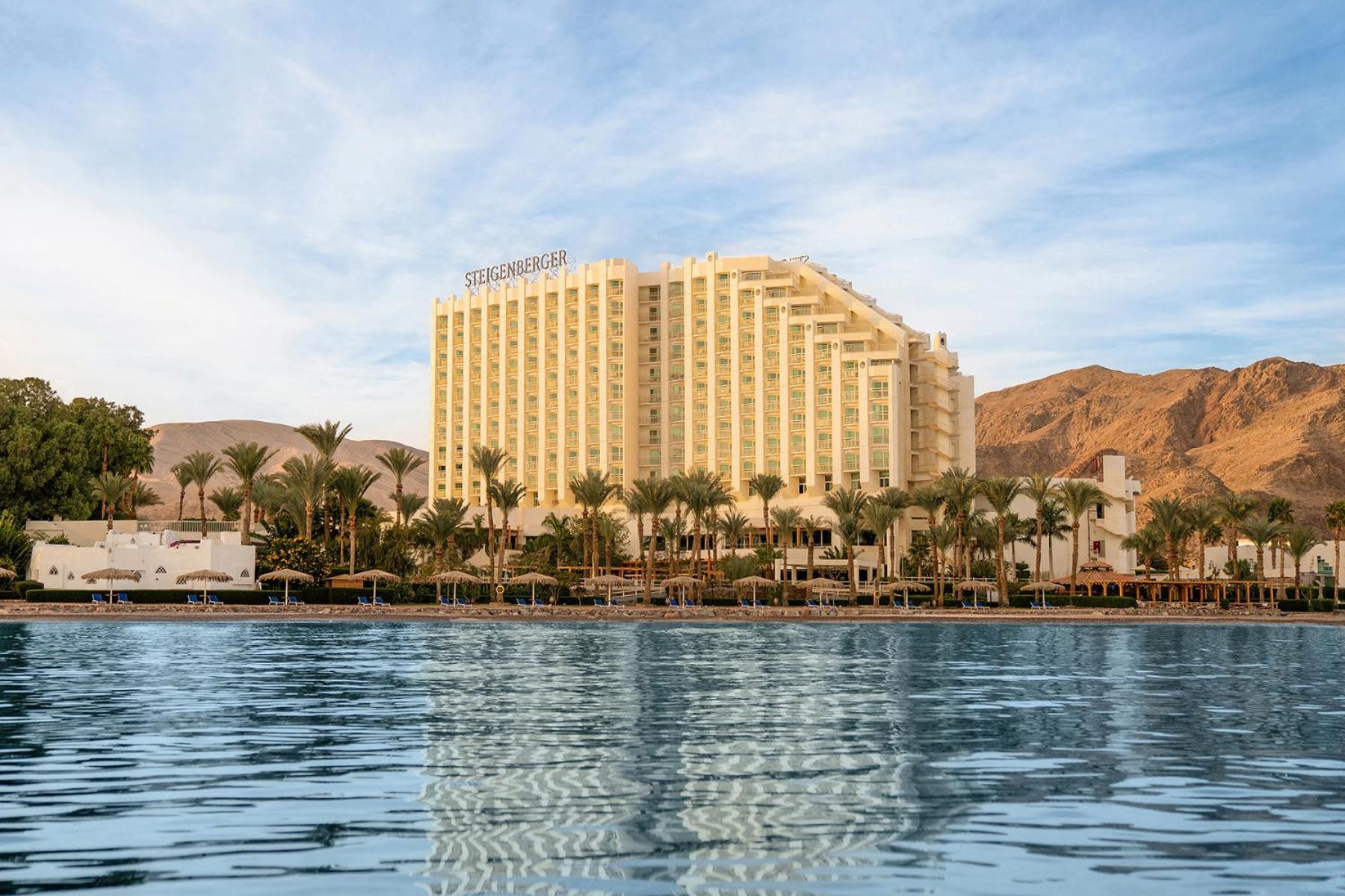 Steigenberger Hotel & Nelson Village, Taba Exterior foto The photo shows a large hotel building next to a body of water, possibly a lake or sea. The hotel is tall and features a modern design with several floors. In the foreground, you can see the calm water reflecting the hotel and the surrounding palm tr