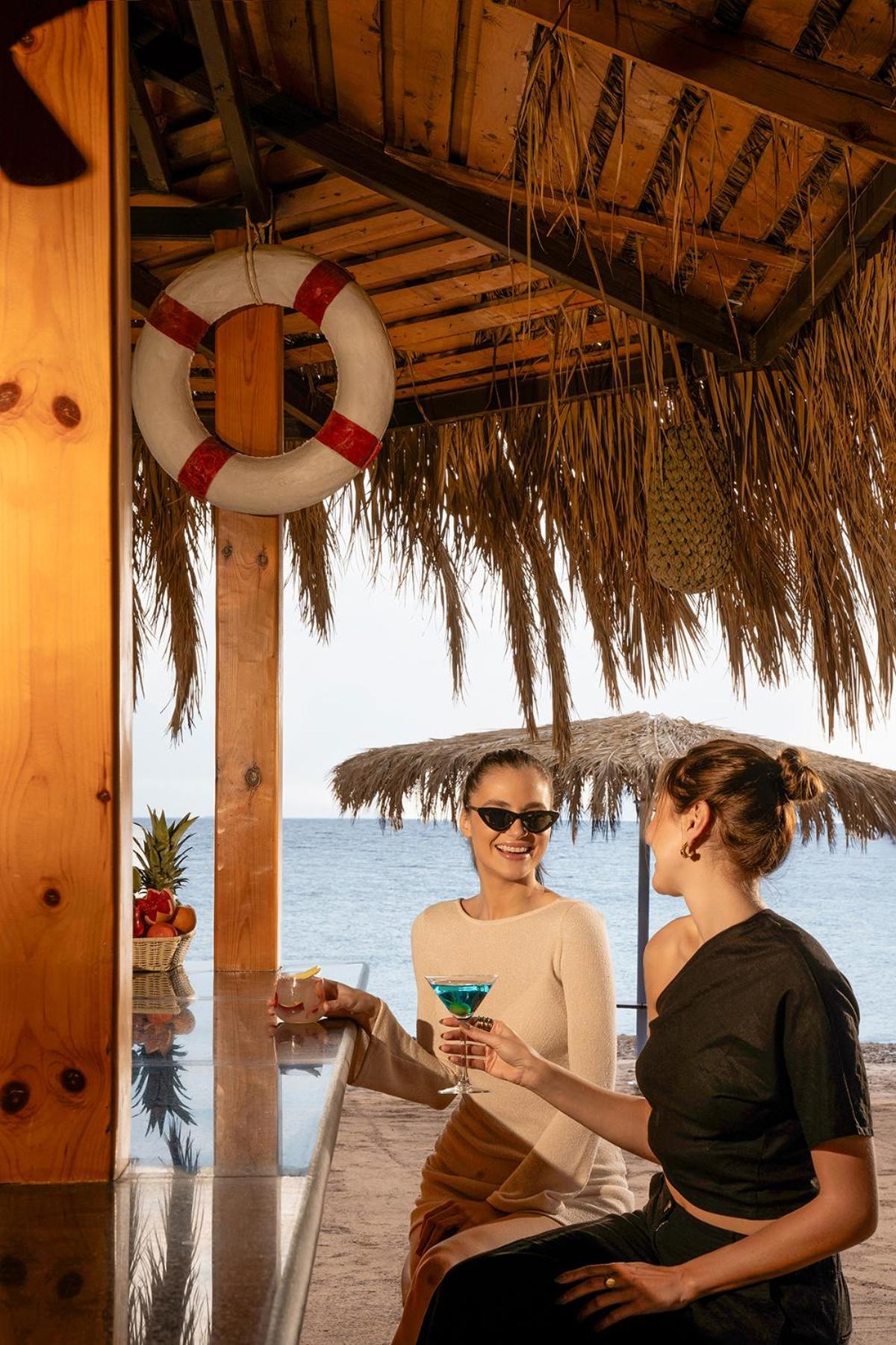 Steigenberger Hotel & Nelson Village, Taba Exterior foto The photo shows two women enjoying a moment at a beach bar. One of the women, wearing sunglasses, is smiling and holding a colorful drink, while the other woman is sitting beside her, wearing a black outfit. The bar is made of wood, and the setting f