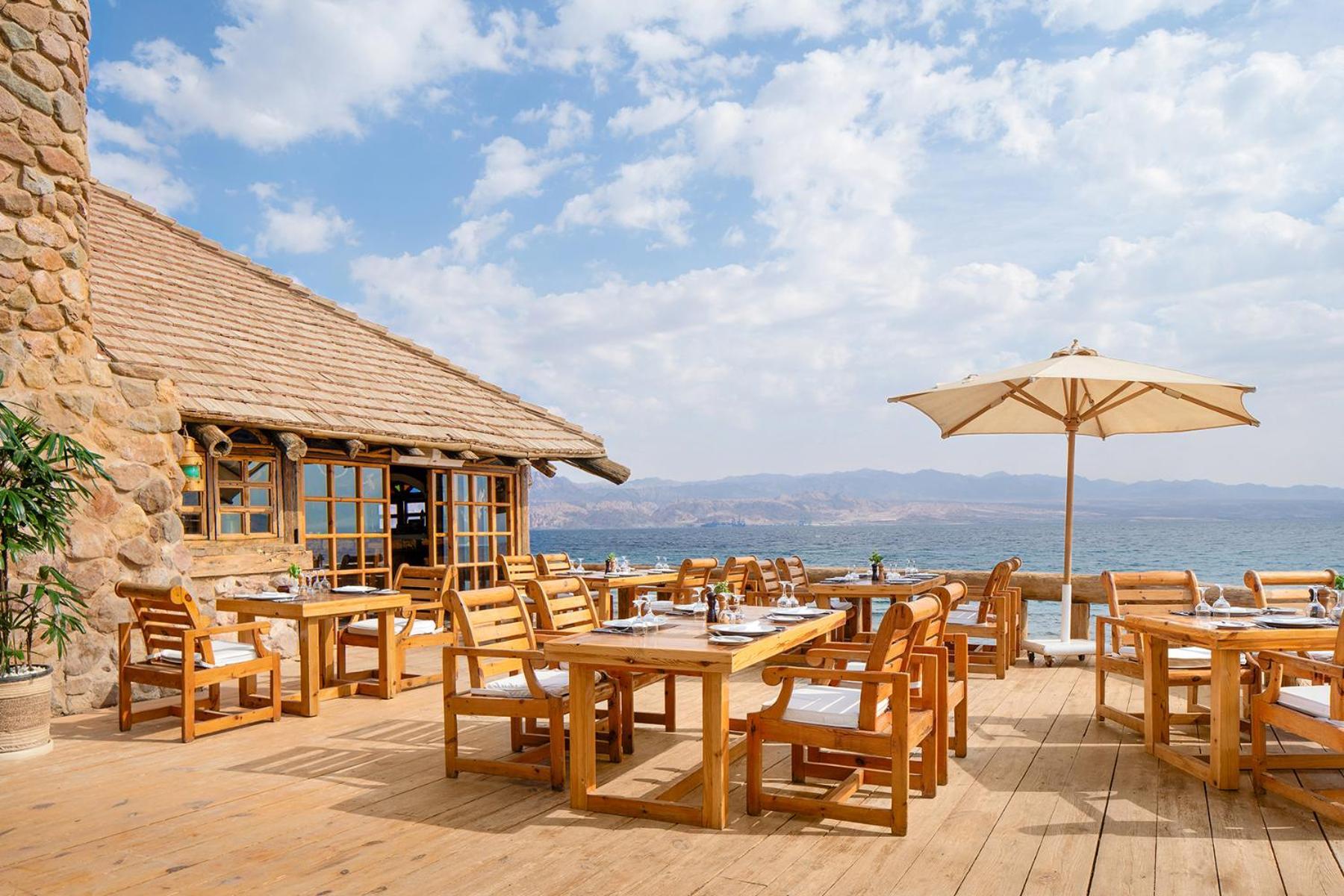 Steigenberger Hotel & Nelson Village, Taba Exterior foto The photo depicts an outdoor dining area at a restaurant. There are several wooden tables and chairs arranged on a wooden deck, along with a large umbrella providing shade for one of the tables. In the background, you can see a building made of stone