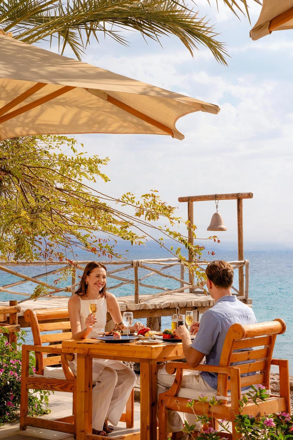 Steigenberger Hotel & Nelson Village, Taba Exterior foto The photo shows a romantic outdoor dining setting by the sea. There are two people seated at a wooden table, enjoying drinks. The woman is smiling, wearing a light-colored dress, while the man, in a blue shirt, appears to be engaged in conversation. 