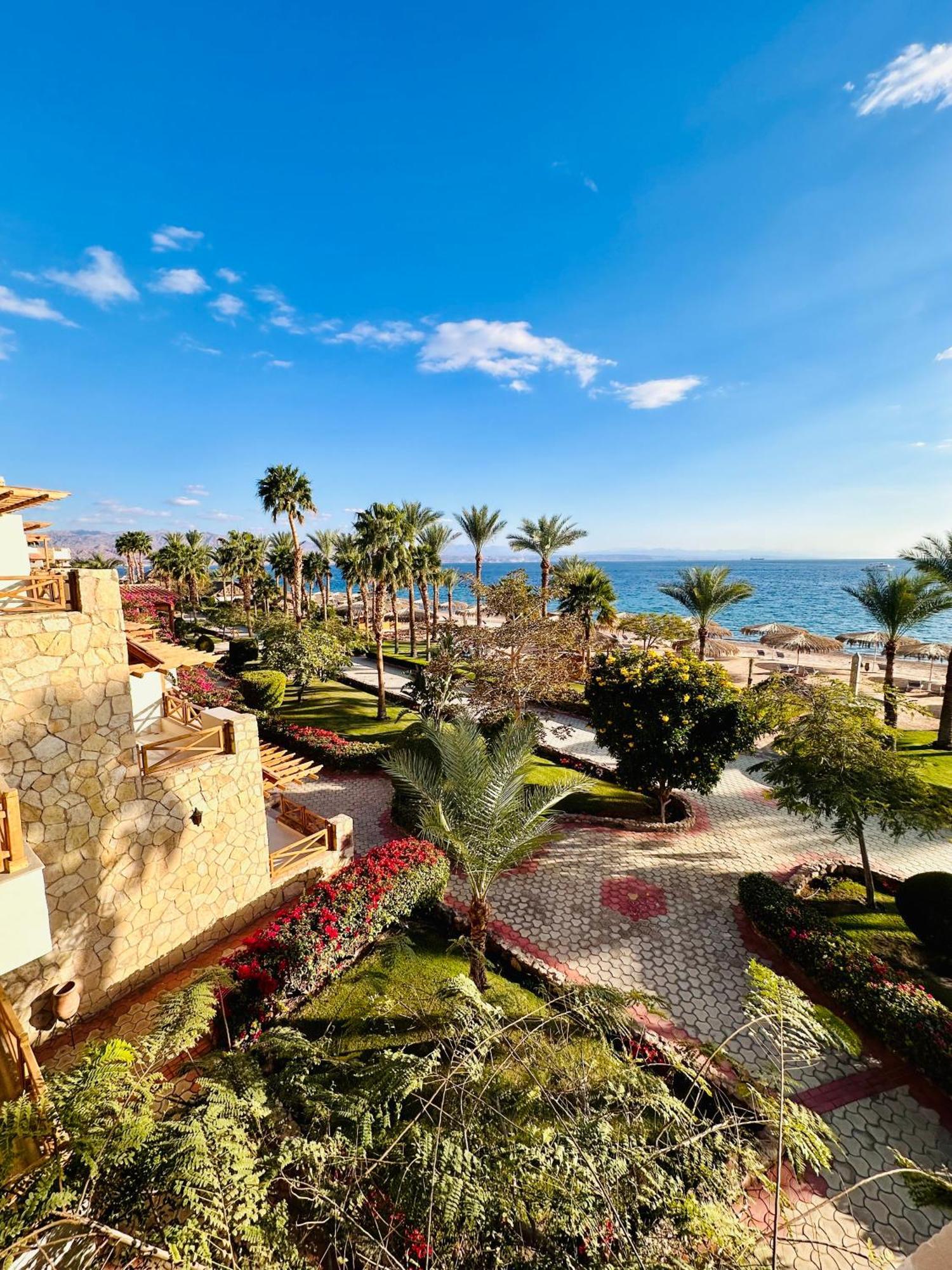 Steigenberger Hotel & Nelson Village, Taba Exterior foto The photo depicts a scenic coastal view featuring a beach surrounded by palm trees and lush greenery. In the foreground, there are manicured gardens and pathways, leading toward the beach area. The clear blue sky and calm sea create a serene atmosphe