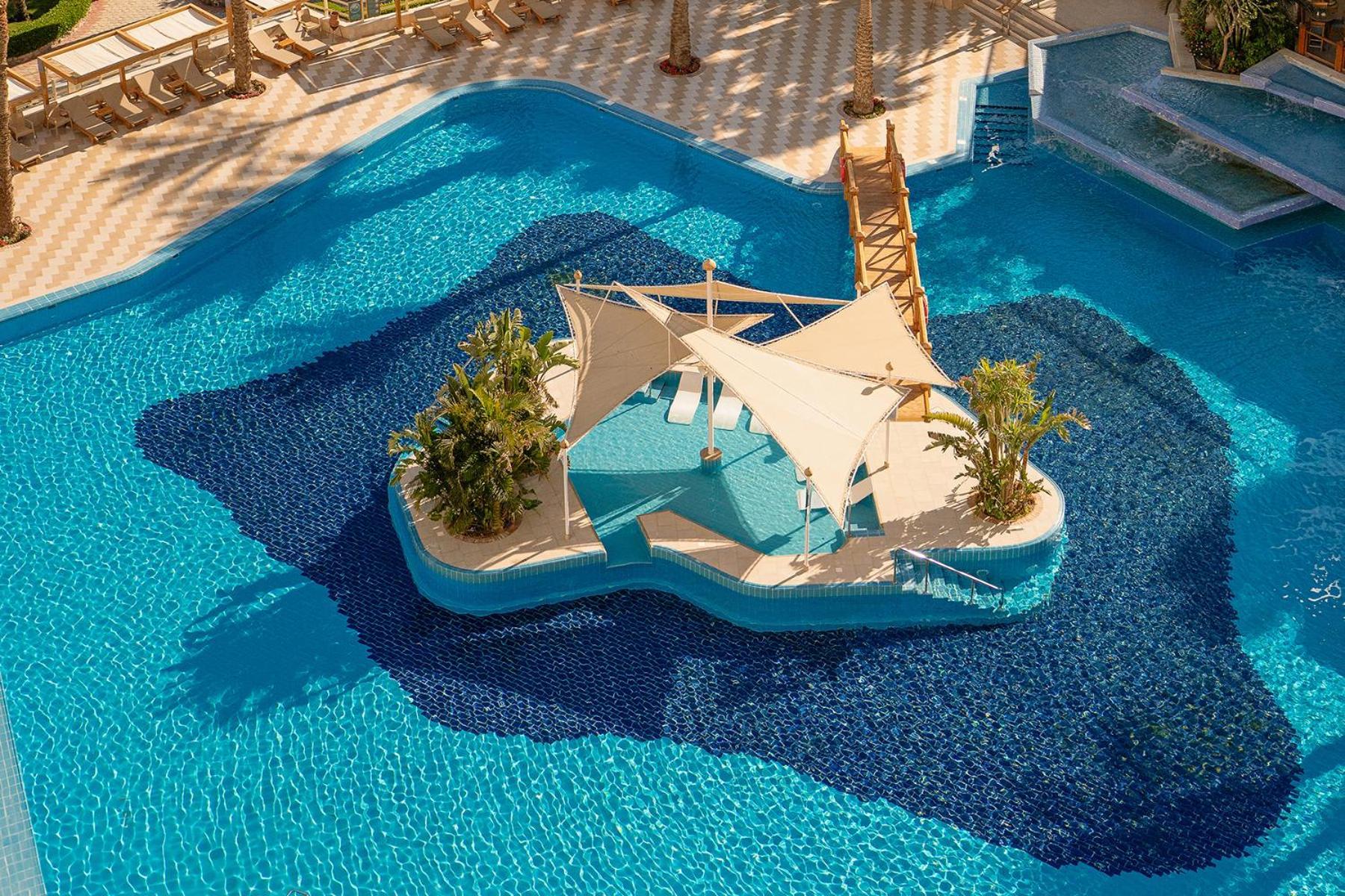 Steigenberger Hotel & Nelson Village, Taba Exterior foto The photo shows a large swimming pool with a vibrant blue color. In the center of the pool, there is an island-like structure featuring a tent with a curved, modern design, providing shade. Surrounding the tent are lush green plants, adding to the tr